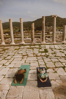 Two individuals practicing yoga on mats in an ancient ruin setting, surrounded by columns and mountains.