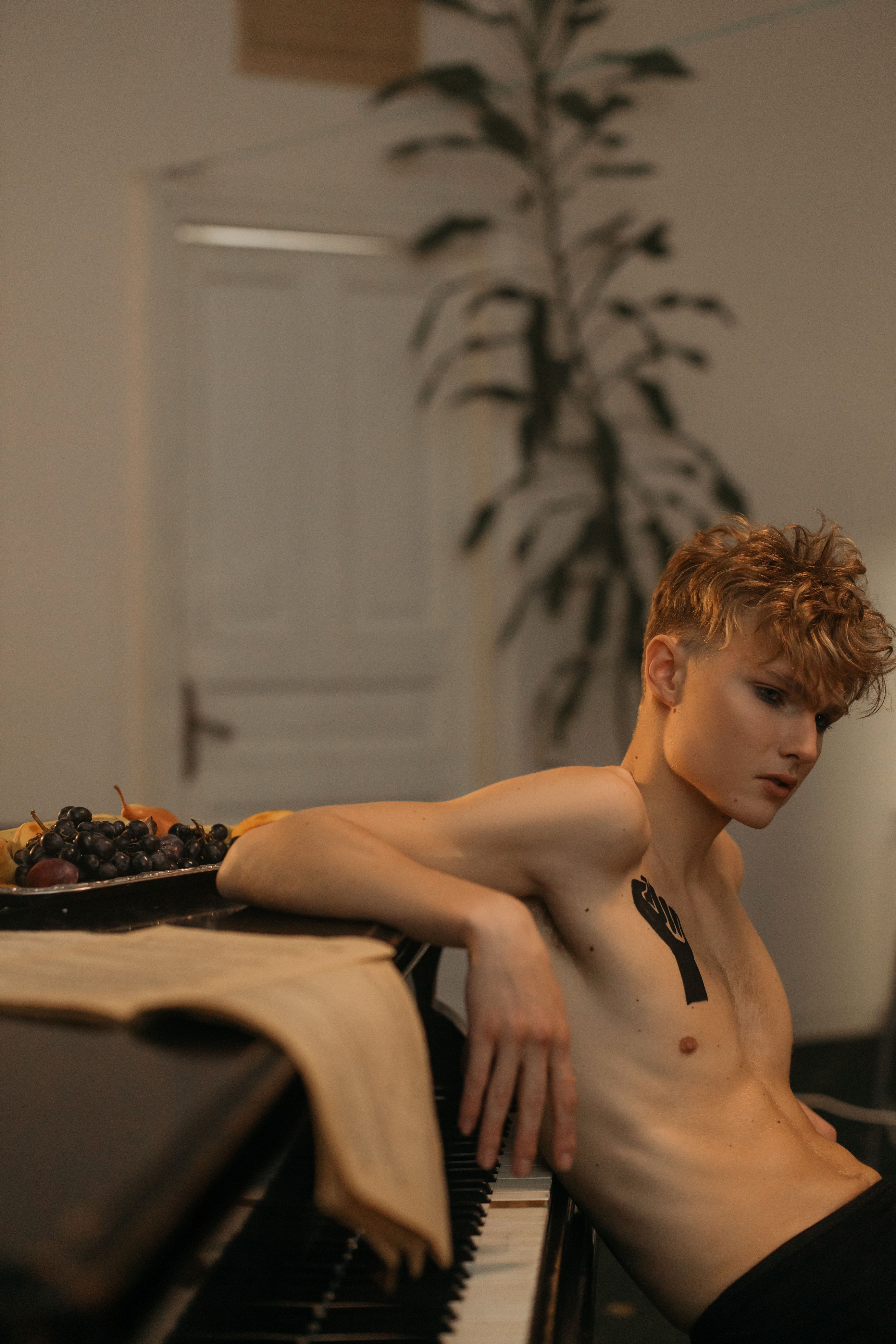 A shirtless man with tattoos leans against a piano in a serene indoor setting, evoking a vintage artistic mood.