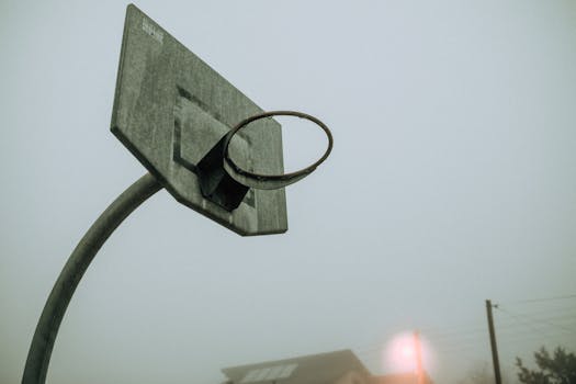 A weathered basketball hoop stands alone in thick fog, creating a moody outdoor scene.