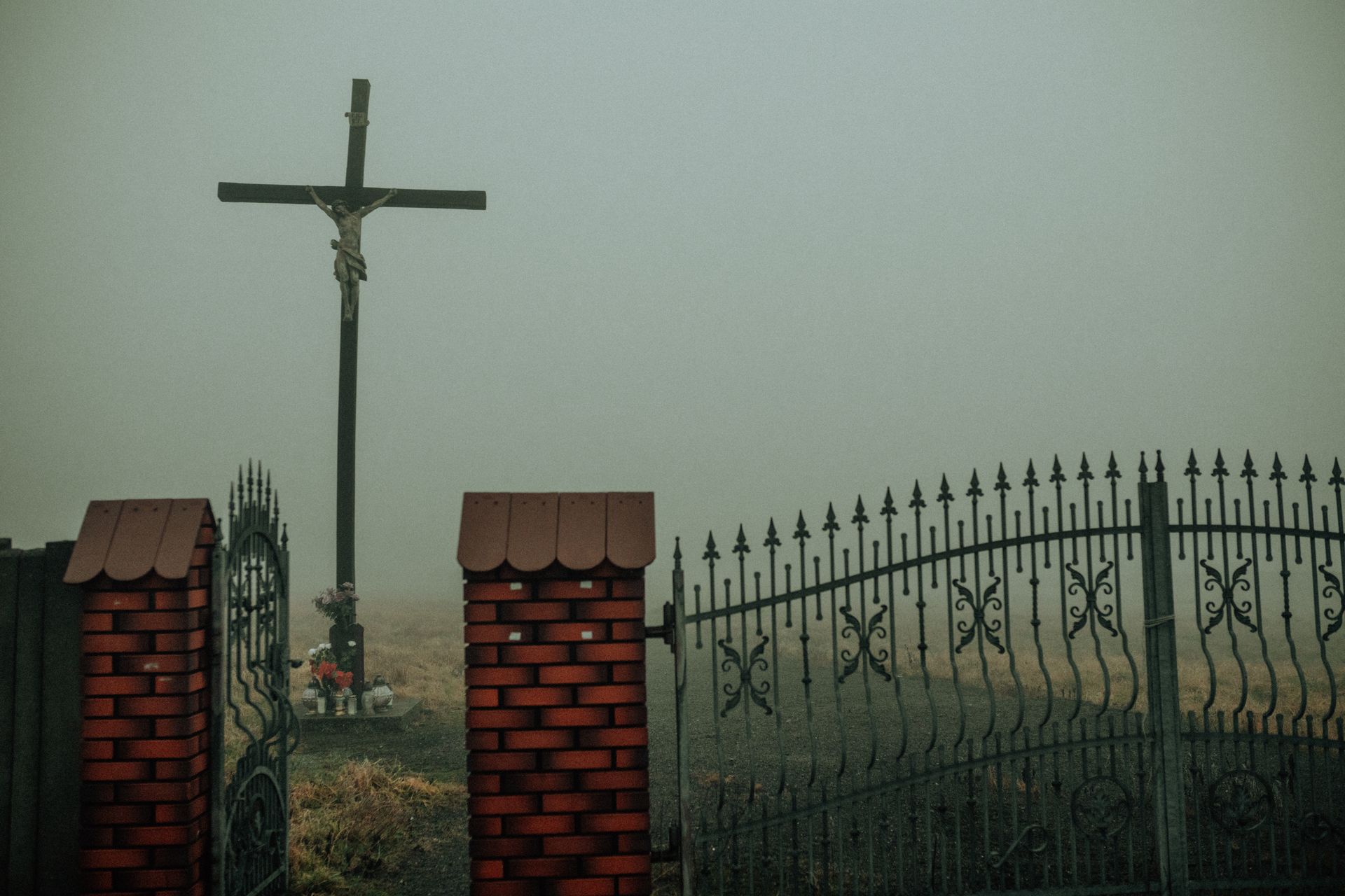 A cross stands behind an intricate gate enveloped in dense fog, creating a somber mood.