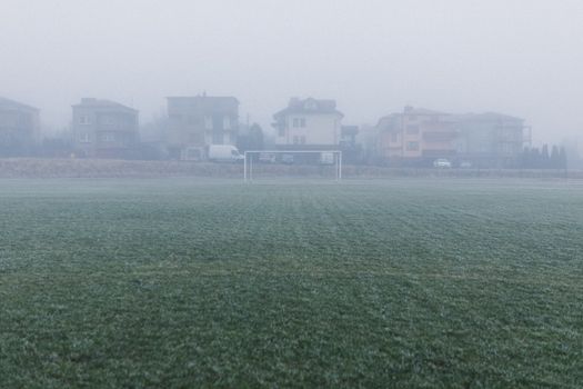 A foggy soccer field with houses in the background creates a serene winter morning scene.