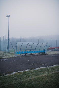 Empty blue seats under a canopy on a fog-covered sports field.