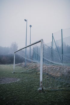 Misty soccer field featuring a weathered goalpost and tall fence under foggy conditions.