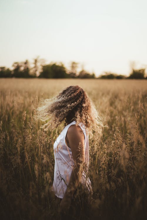 Side view of unrecognizable female with flying hair wearing white t shirt standing in meadow with tall plants in nature on blurred background