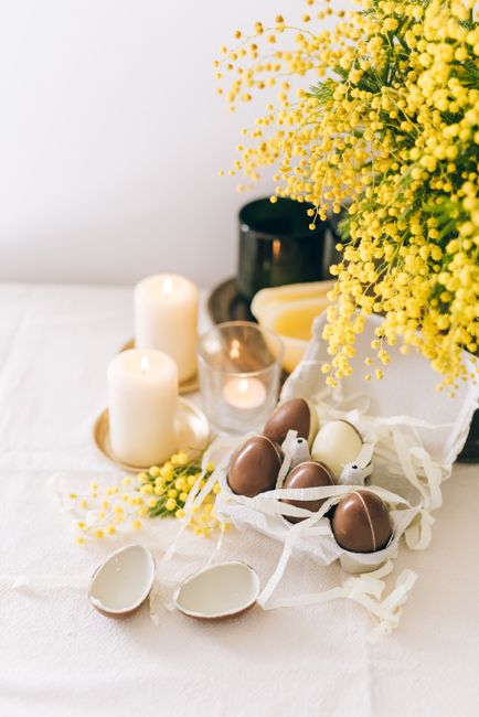 Elegant Easter-themed arrangement with chocolate eggs, candles, and mimosa flowers on a table.