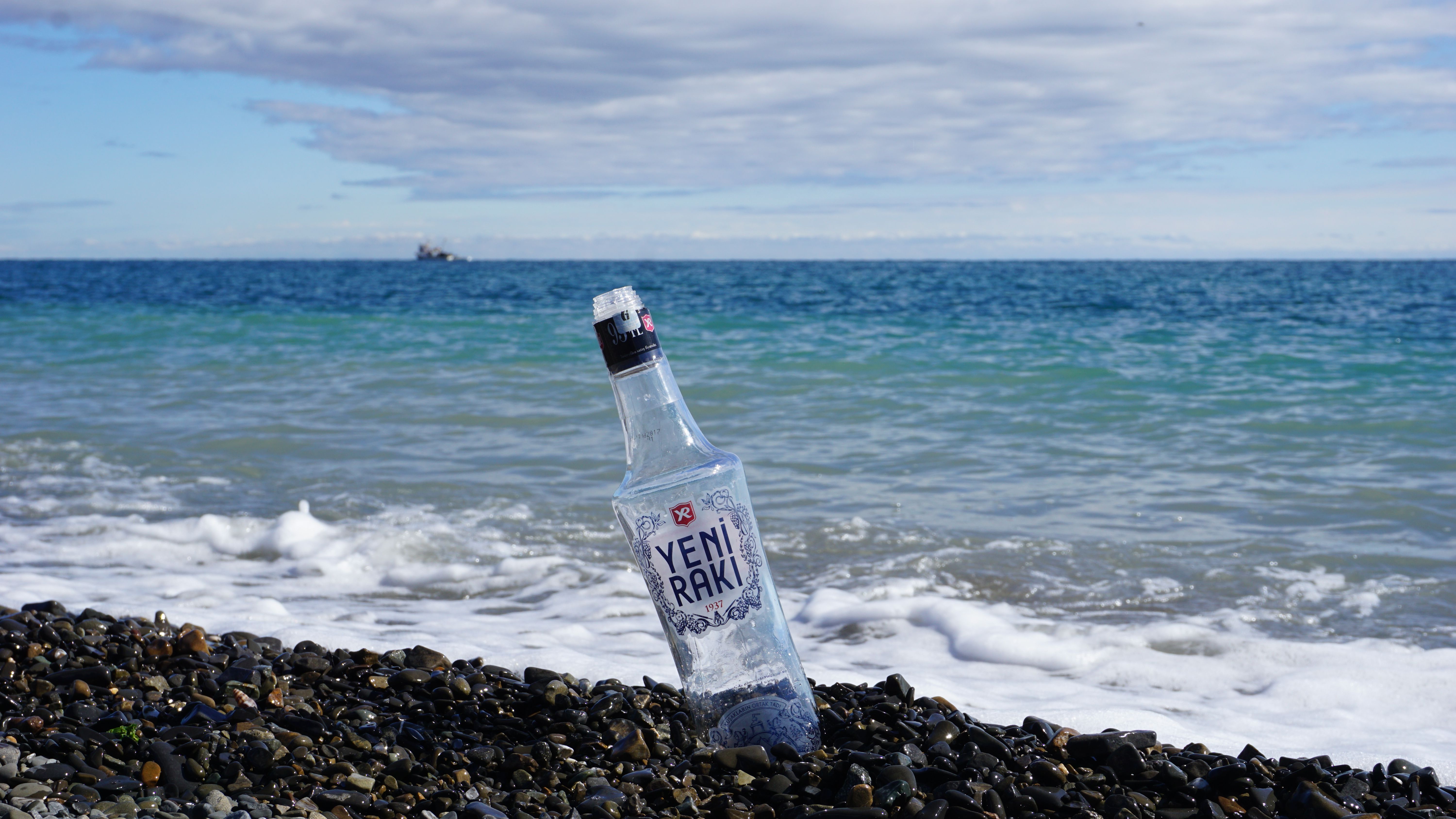 Clear Yeni Rakı bottle on a pebbly beach in Şarköy, Türkiye, with waves gently crashing on the shore.