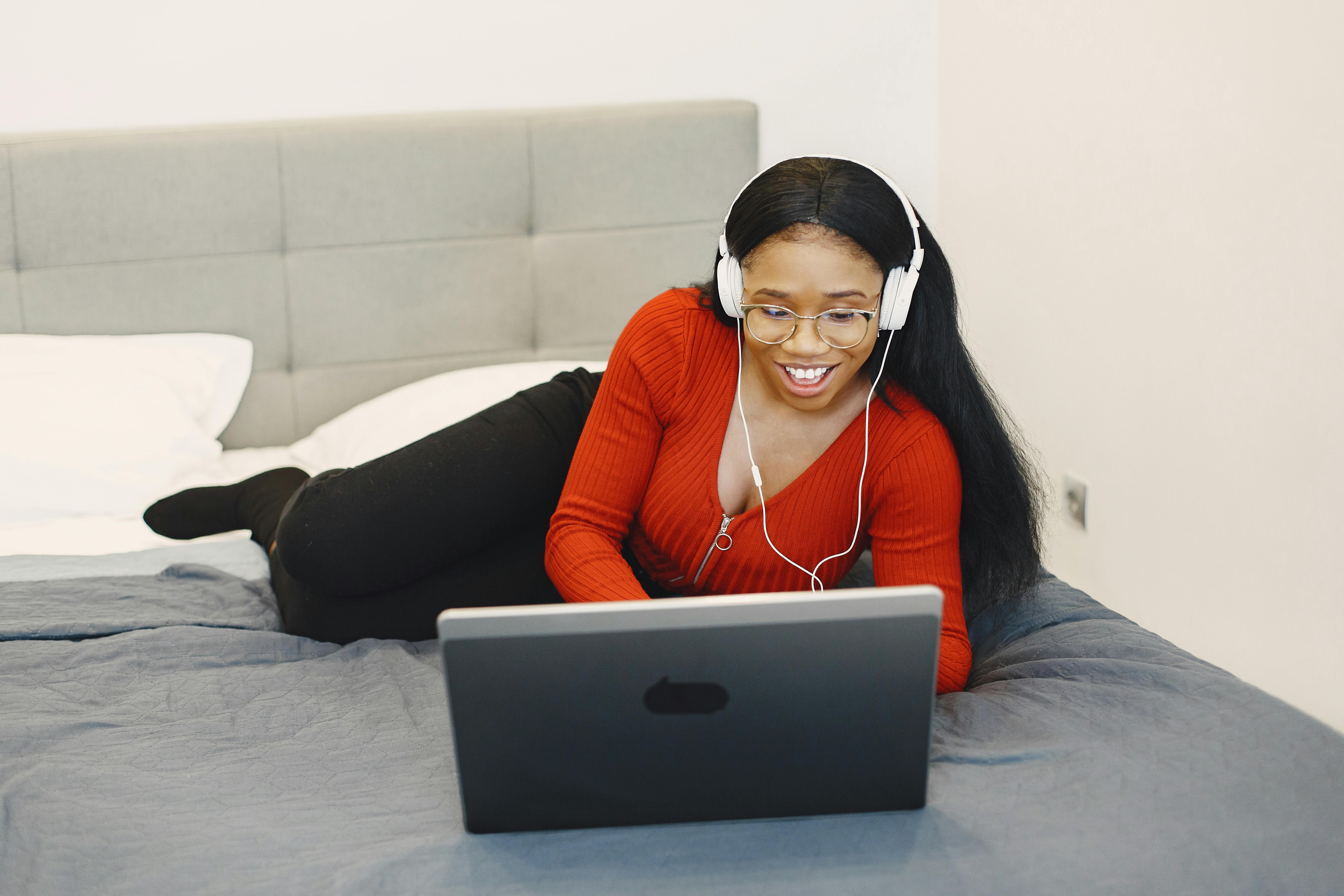 A cheerful young woman with headphones using a laptop while relaxing on her bed indoors.