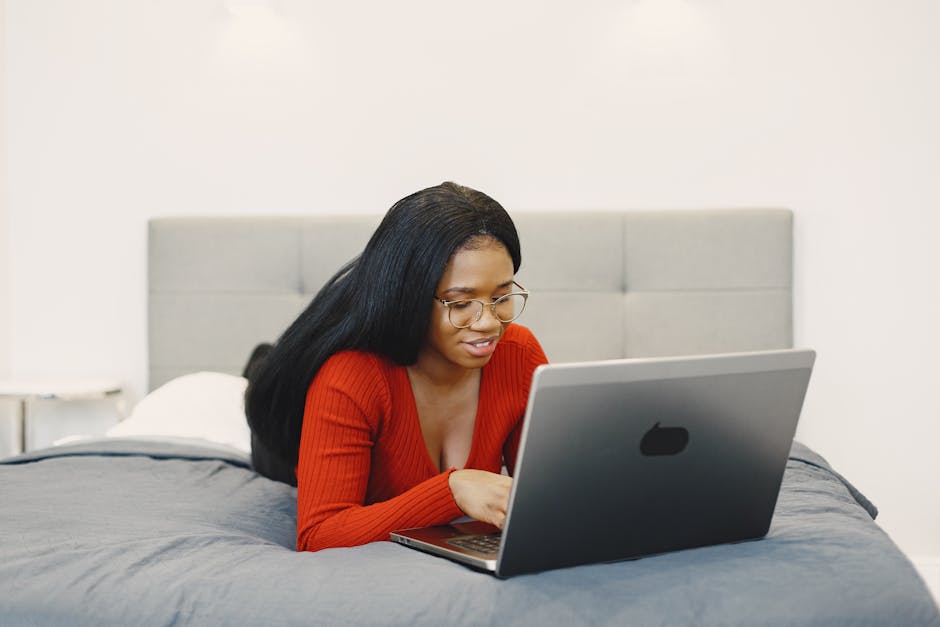 Stop Guessing Mattress Firmness—Use This Weight Chart Young woman with glasses working on a laptop while lying on a bed indoors.