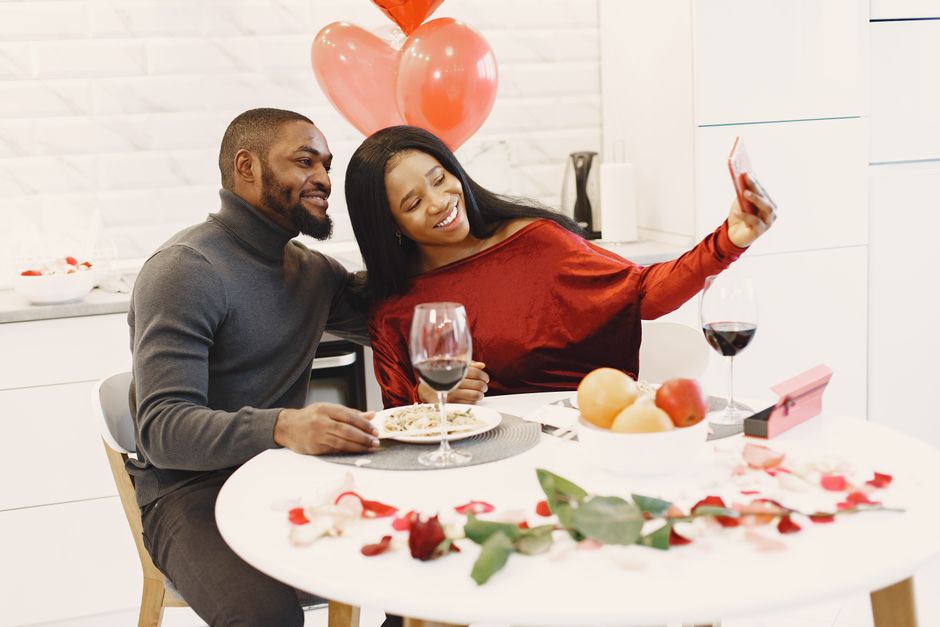 Couple enjoying a romantic dinner with red wine and balloons, taking a selfie for a special occasion.