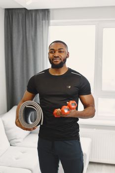 Man in a t-shirt holding dumbbells and yoga mat indoors, ready for a workout.