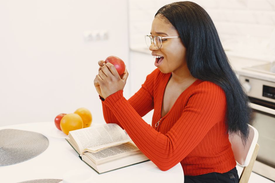 Black woman in red top holds apple, reading a book at a kitchen table.