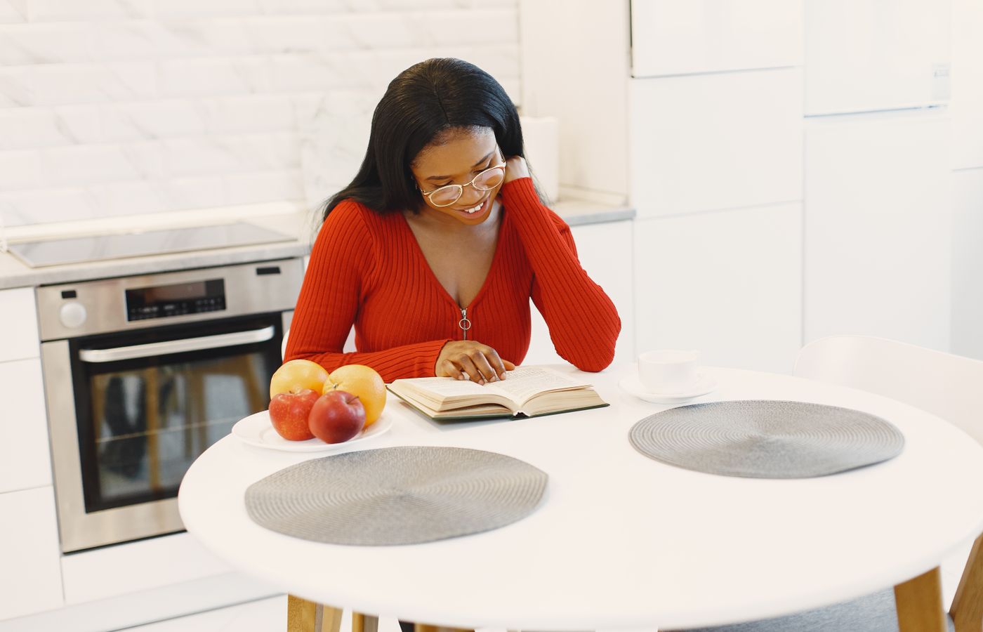 A woman in a red sweater reading at a round table in a bright modern kitchen.