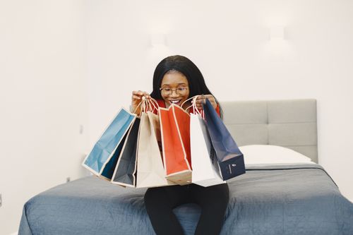 Free Happy woman holding colorful shopping bags, seated on a bed in a cozy bedroom. Stock Photo