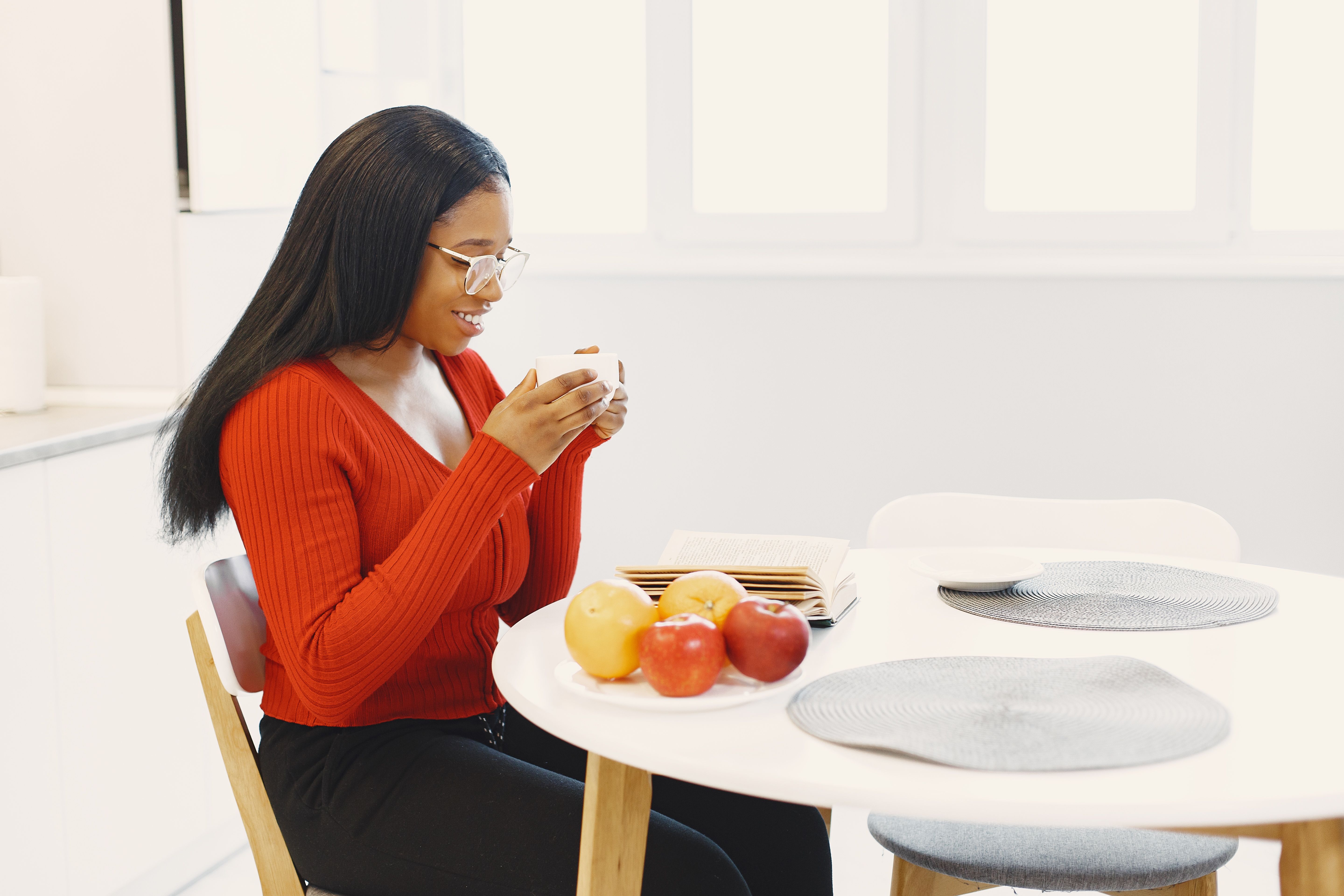A woman with eyeglasses enjoys coffee while reading at a modern kitchen table with fresh fruit.