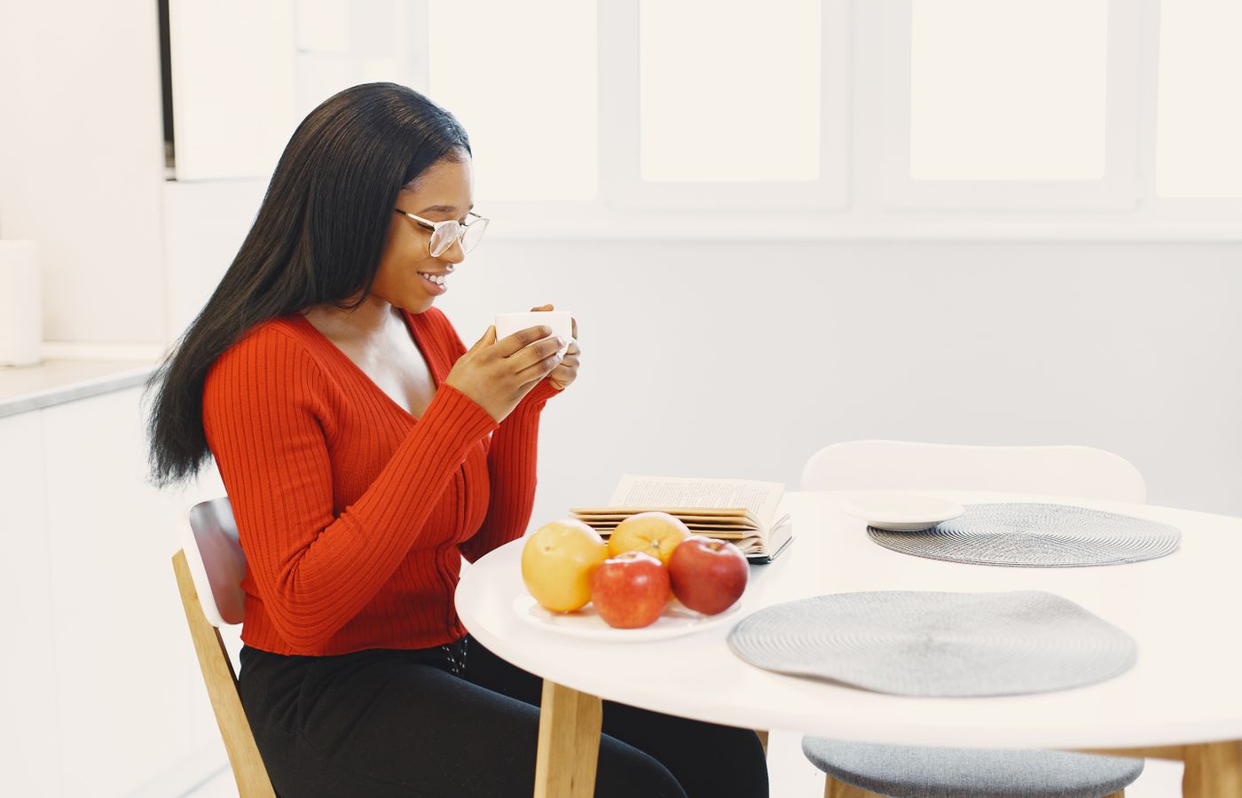 A woman with eyeglasses enjoys coffee while reading at a modern kitchen table with fresh fruit.
