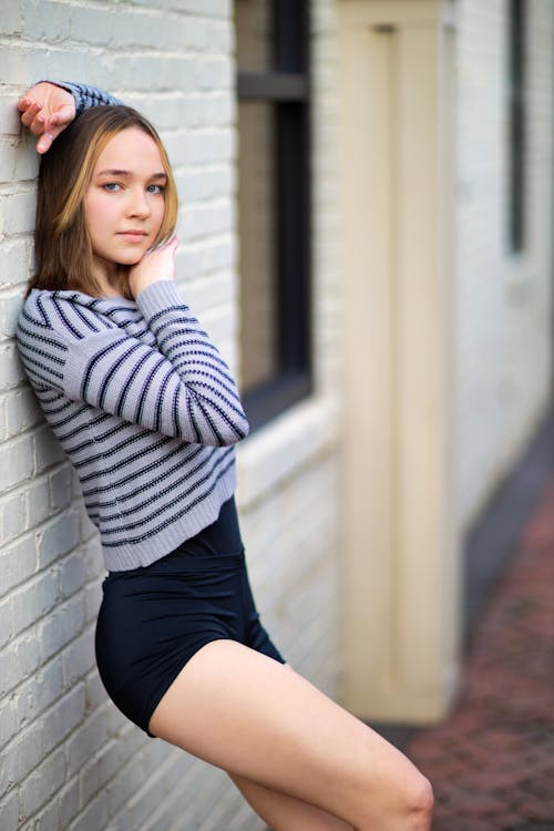 Side view beautiful young slim female in shorts leaning on brick building wall with arm raised and looking at camera calmly
