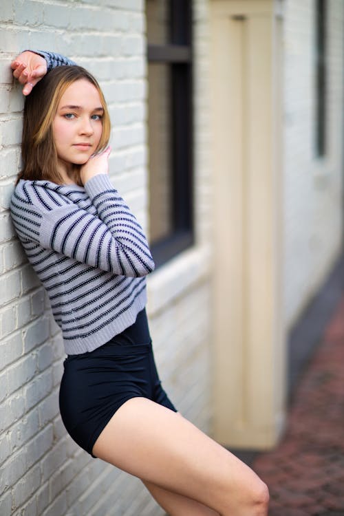 Free Side view beautiful young slim female in shorts leaning on brick building wall with arm raised and looking at camera calmly Stock Photo