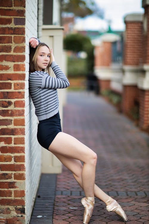 Side view full body fit sporty female in shorts and pointe shoes standing gracefully on tiptoes against brick building while touching head and looking at camera