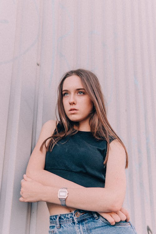 Low angle of serious young female in casual clothes standing with crossed arms and looking away