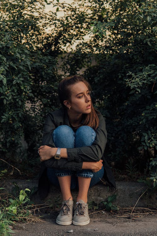 Full body of upset female embracing knees sitting on curb near green bushes and thoughtfully looking away