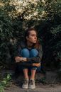 Full body of sad female embracing knees sitting on ground among bushes with green foliage