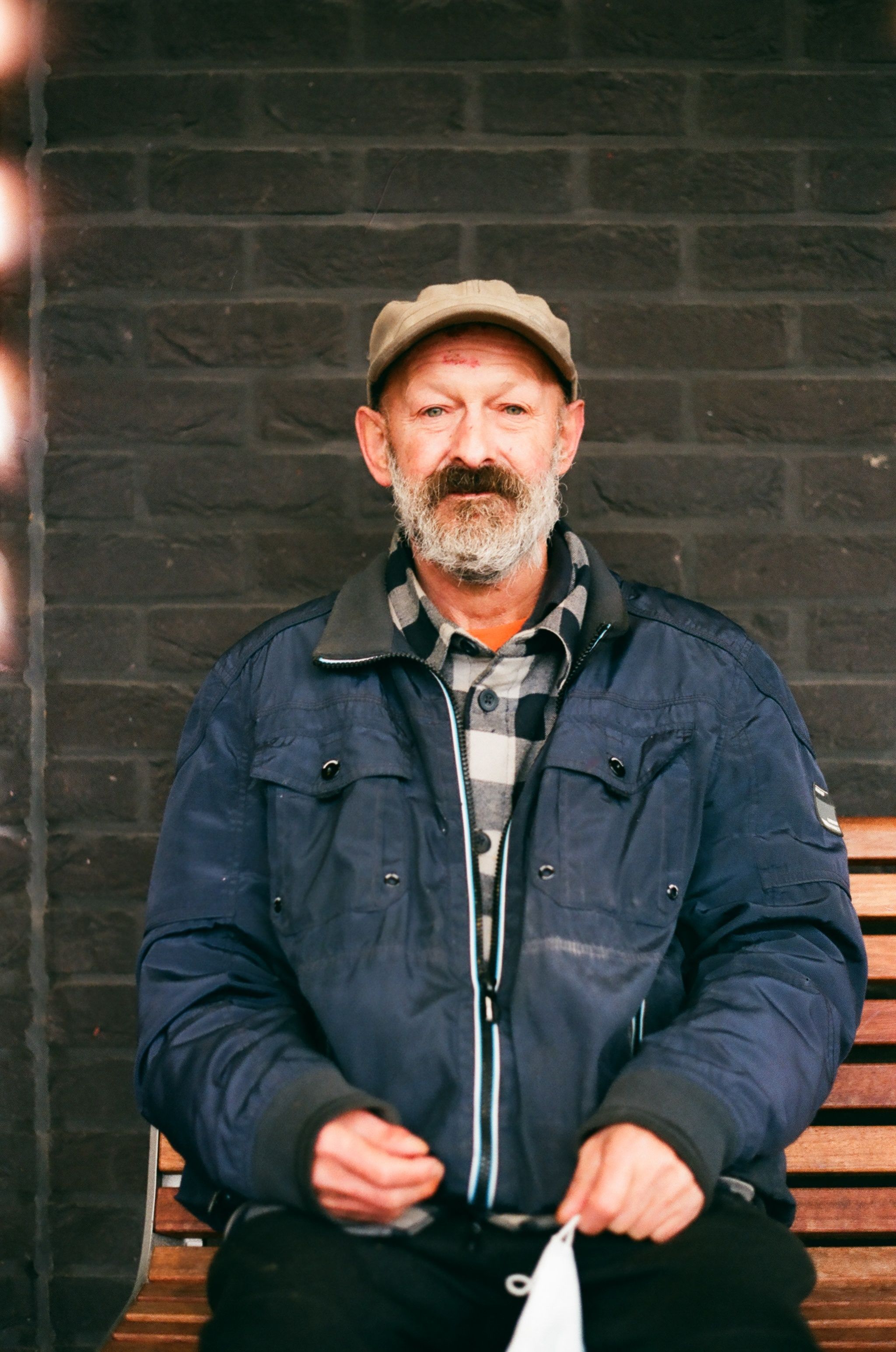Elderly man with a beard and cap sitting on a wooden bench against a brick wall.