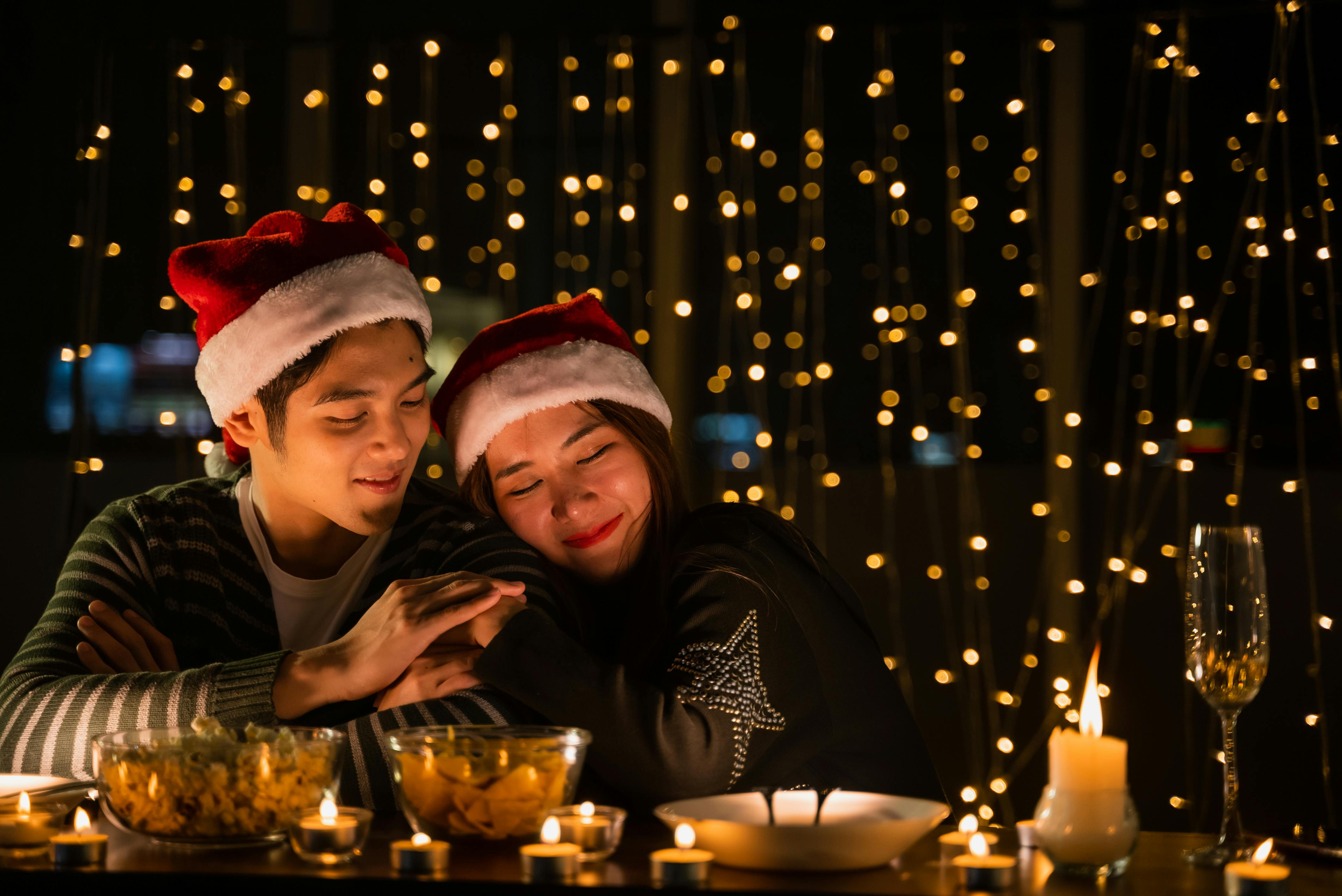 A couple enjoying a cozy Christmas dinner illuminated by candles and string lights.