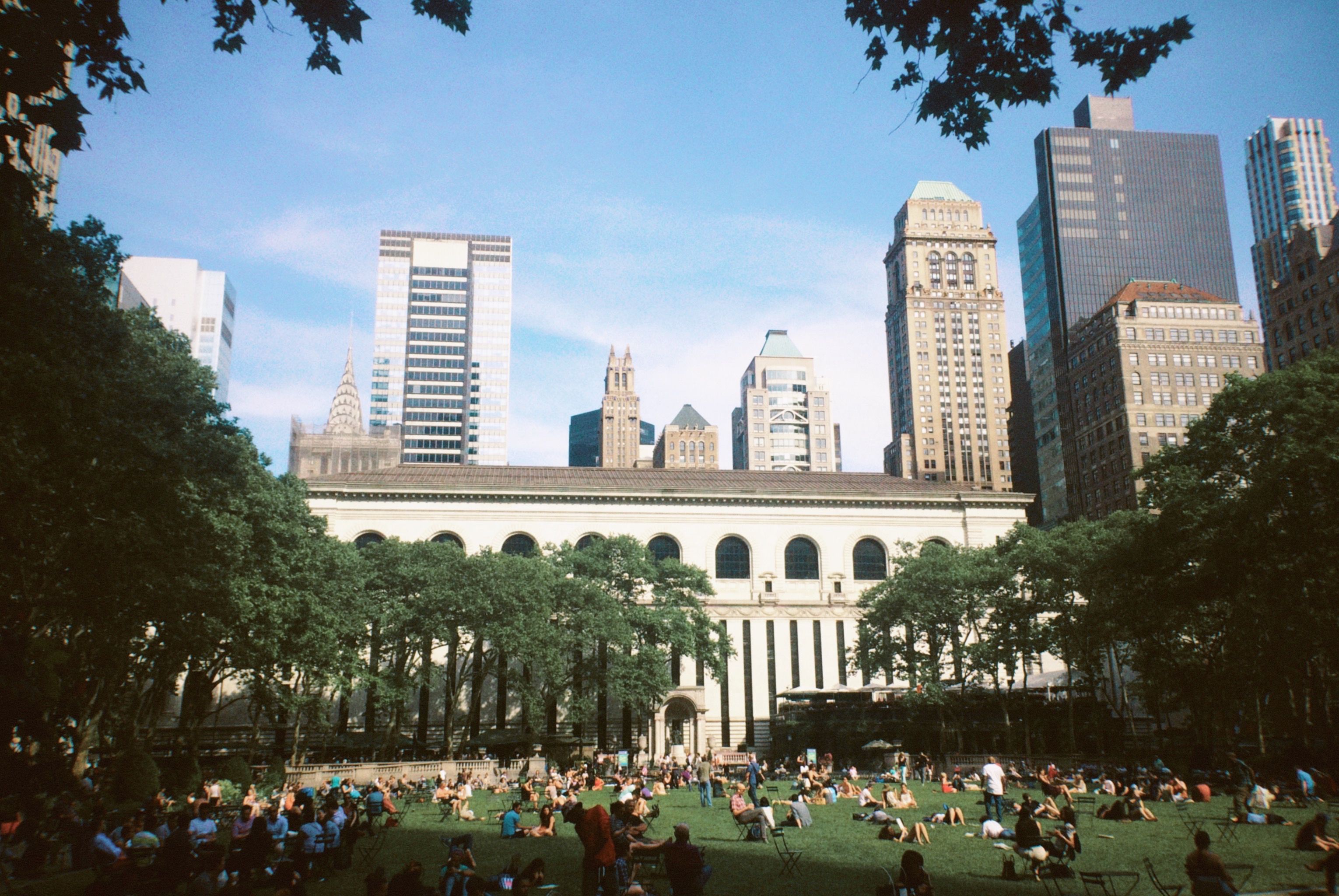 People sitting on a sunlit lawn in front of a long stone library building with arched windows, with skyscrapers rising behind it