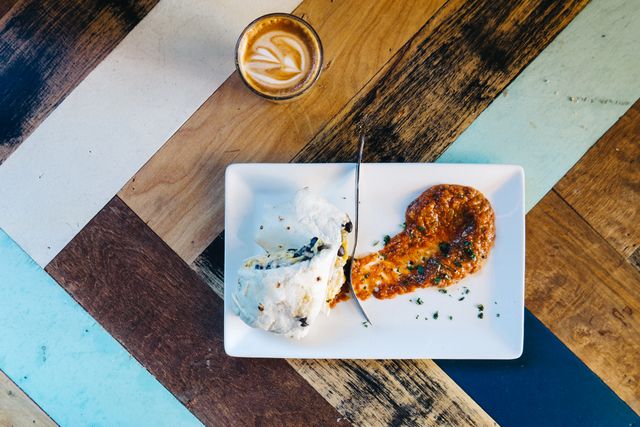Burrito with sauce and coffee on a colorful wooden table, viewed from above.