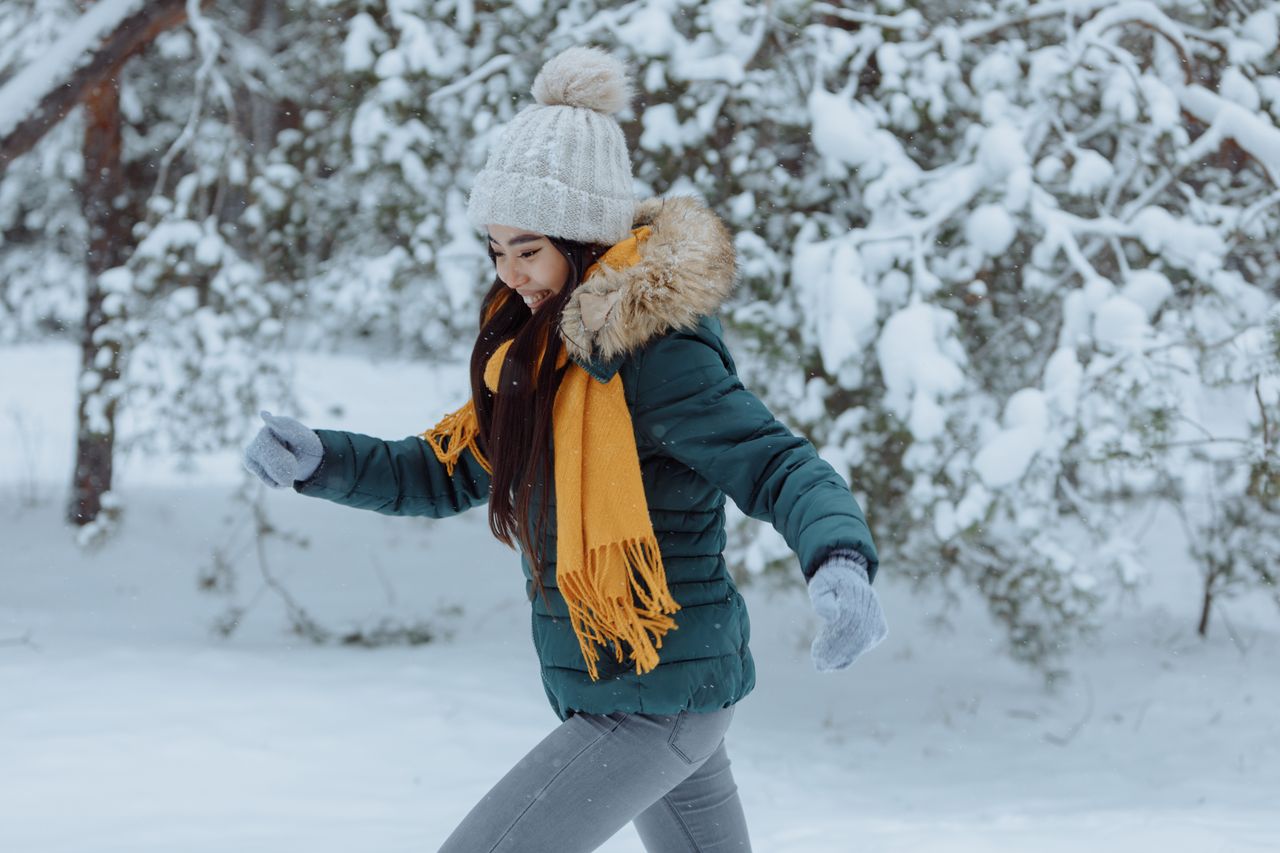 Woman in a green puffer jacket, yellow scarf and white toque jogging through a snowy forest
