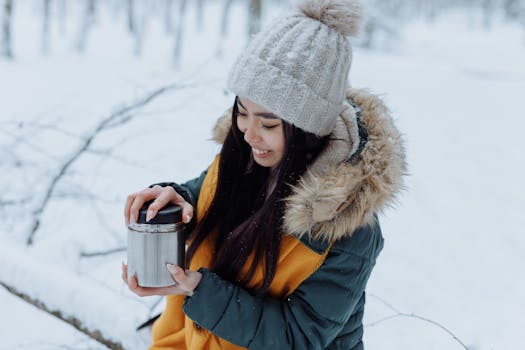 Asian woman in winter attire holding a thermos, smiling amidst a snowy landscape.