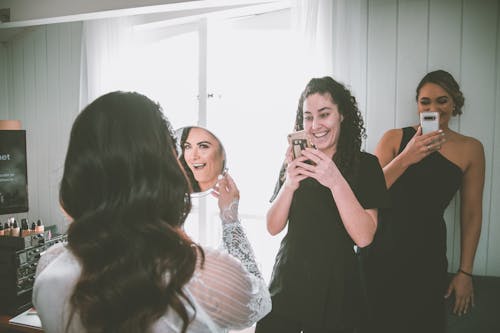 Astonished young bride looking in mirror near positive bridesmaids taking photos on smartphone