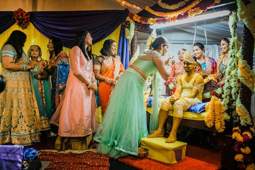 Free Happy Indian groom sitting on couch in decorated room with women during traditional wedding ritual Stock Photo