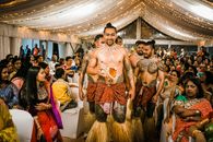 Young shirtless Samoan male dancers in traditional outfits standing amidst Indian guests in decorated canopy tent during wedding party