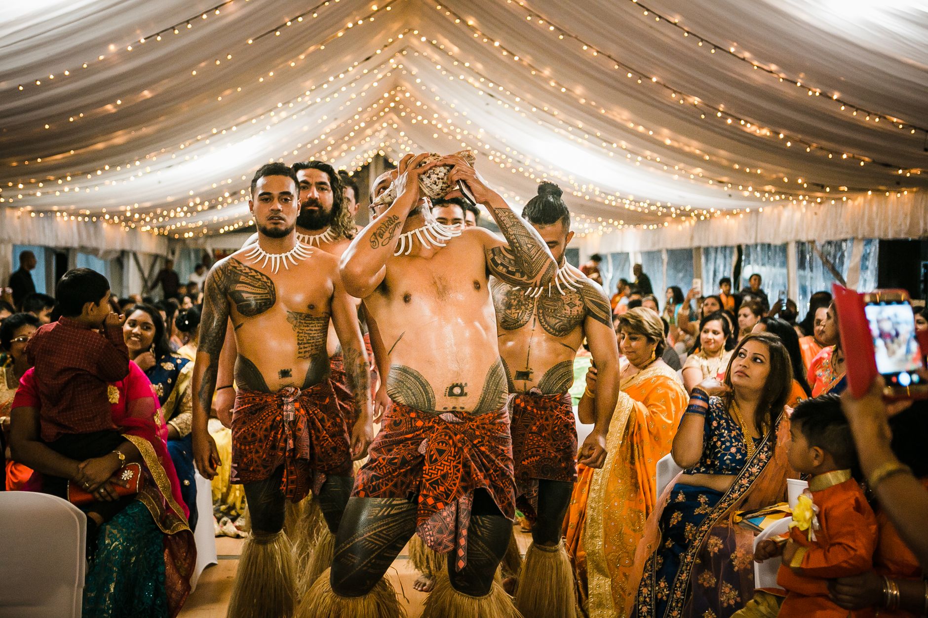 Young tattooed shirtless Samoan men performing dance in decorated hall amidst multigenerational guests during traditional Indian wedding