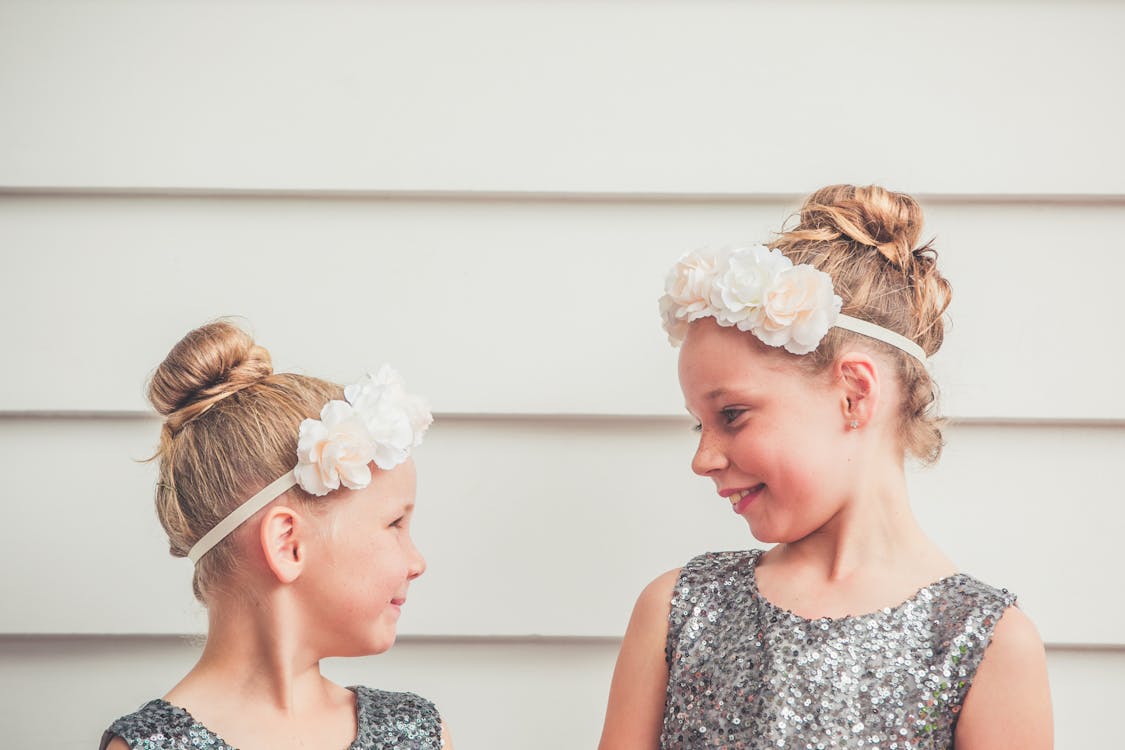 Cheerful little adorable sisters in silver dresses and white headbands smiling and looking at each other