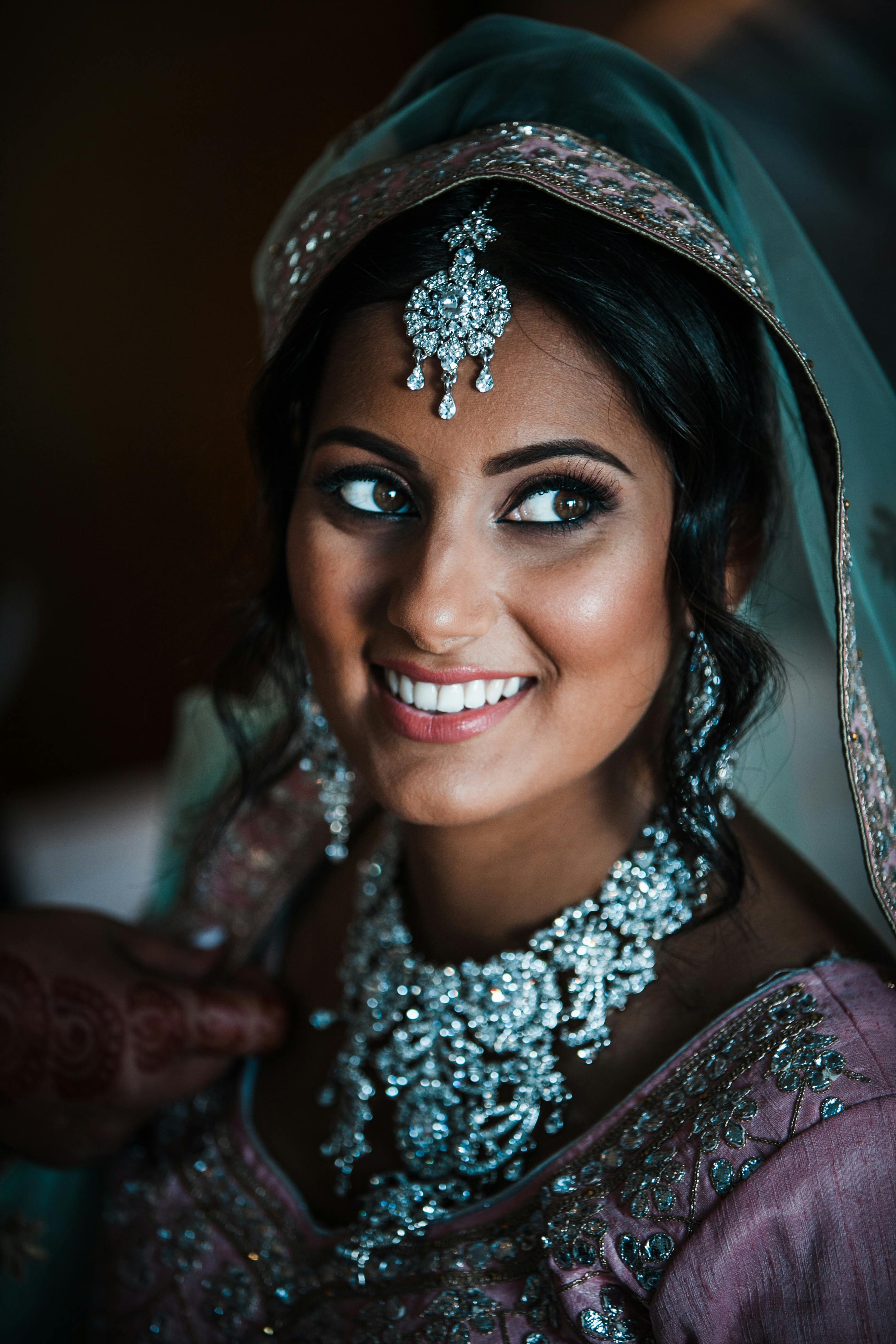 Positive Indian bride wearing traditional maang tikka accessory and necklace looking up while sitting in room in authentic garment during wedding celebration on blurred background