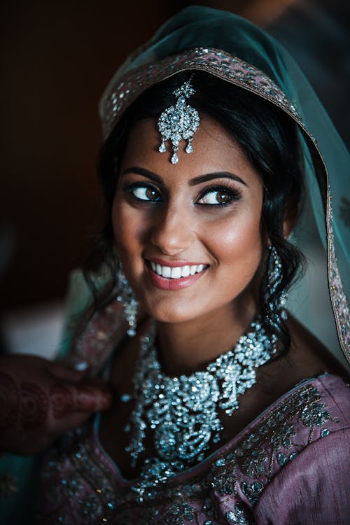 Positive Indian bride wearing traditional maang tikka accessory and necklace looking up while sitting in room in authentic garment during wedding celebration on blurred background