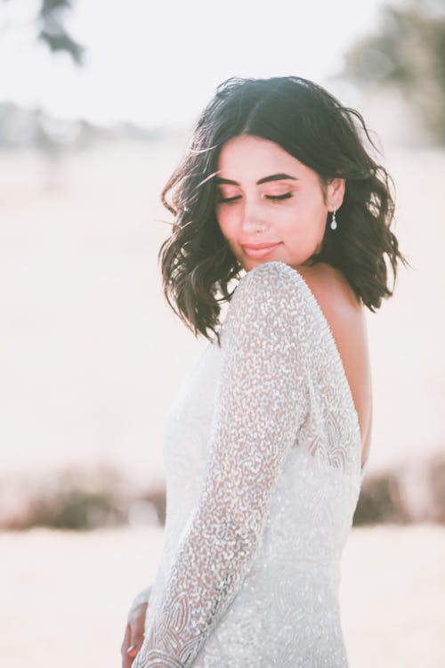 Charming female in elegant white dress standing against blurred nature in daytime