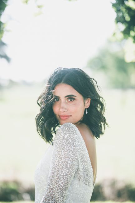 Calm woman with dark hair in elegant stylish white dress standing among green plants in nature and looking at camera in daytime