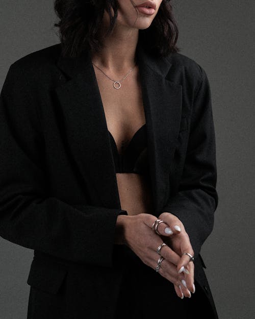 Crop unrecognizable female wearing black classy blazer on underwear with silver rings and necklace against gray background