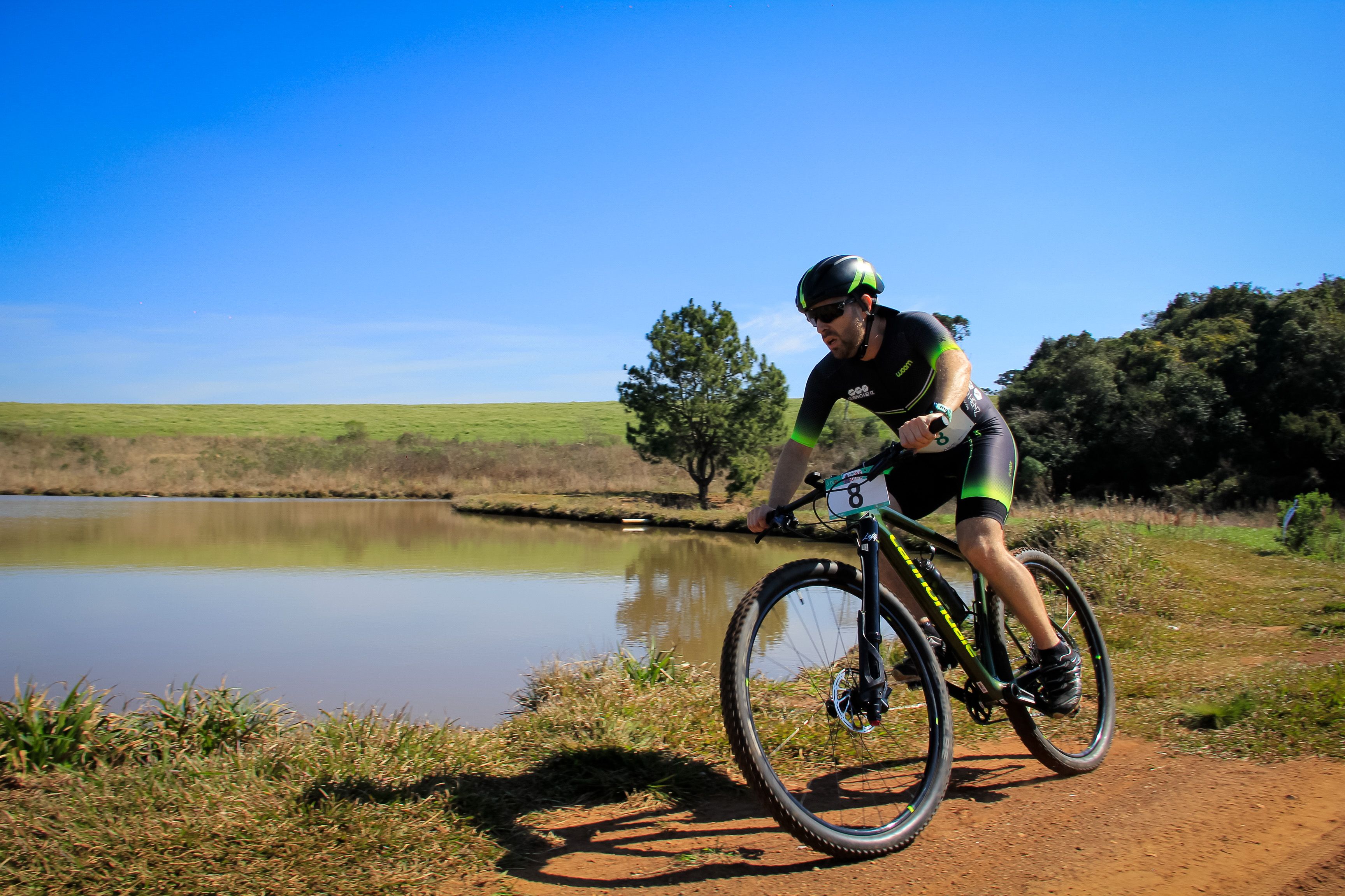 Cyclist racing on a dirt trail near a lake under clear blue skies.