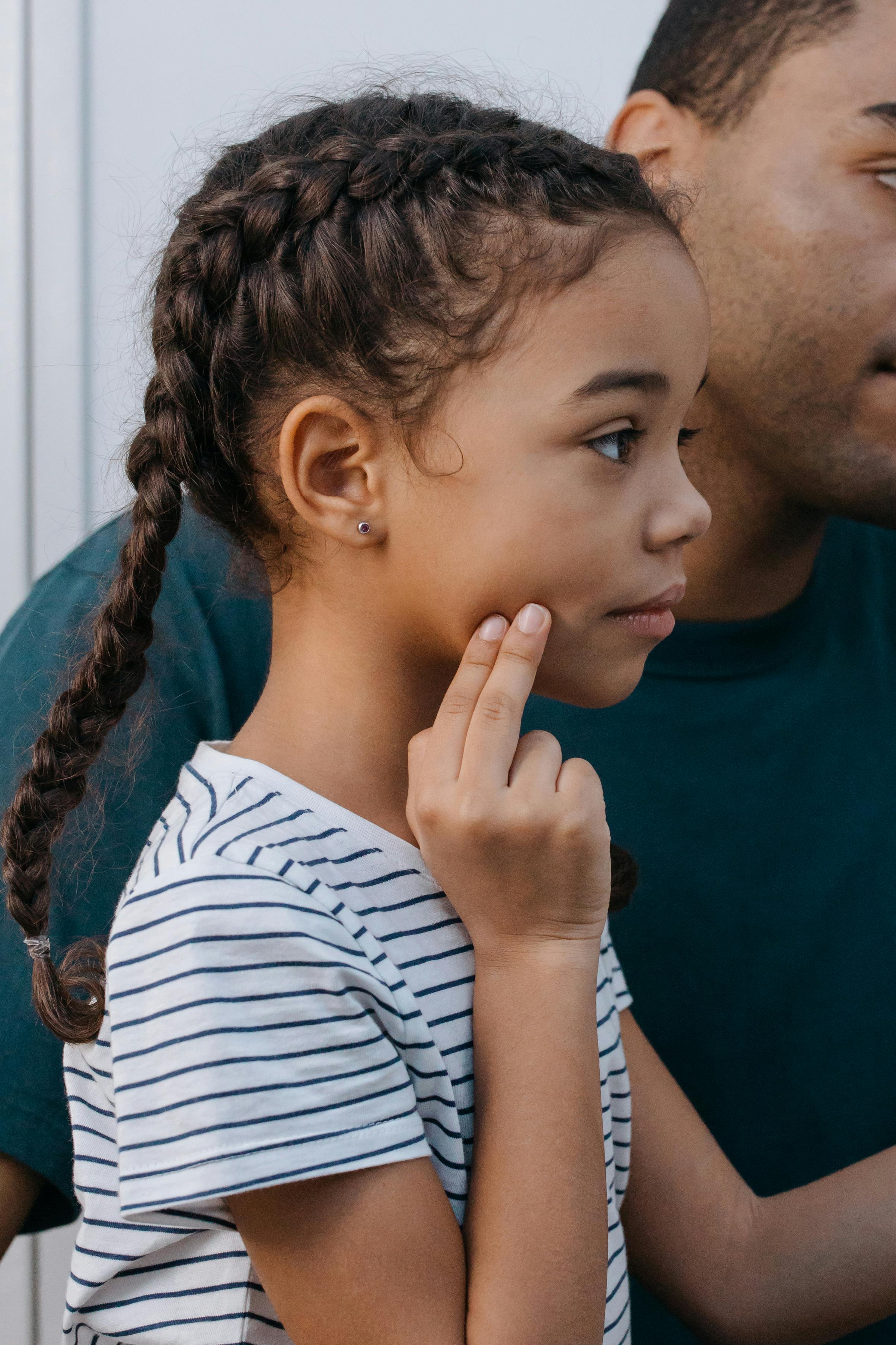 A young girl with braids touching her cheek, expressing toothache discomfort indoors.