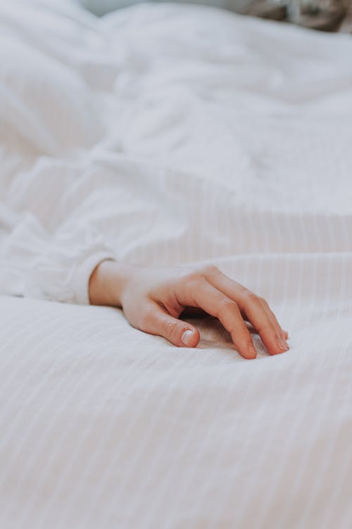 Crop hand of unrecognizable person in white shirt lying on comfortable soft bed in morning