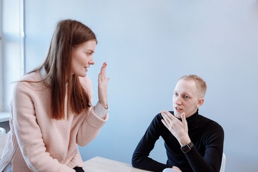 A young man and woman engage in a casual conversation indoors with a light pastel background.
