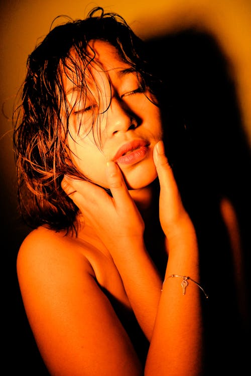 Young sensual ethnic female with wet hair touching face with closed eyes under light