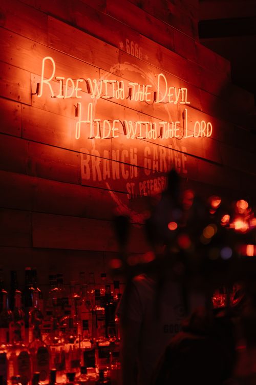 Free Cozy bar interior with neon sign 'Ride with the Devil, Hide with the Lord' above illuminated bottles. Stock Photo