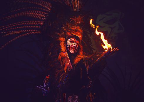 A striking fire performer in vibrant traditional costume with face paint and feather headdress.