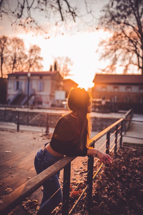 Free Side view of anonymous female leaning on urban fence while admiring residential houses under shiny sky at sundown Stock Photo