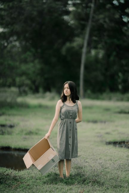 Young contemplative female in sundress with hand behind back and empty carton box looking away on lawn